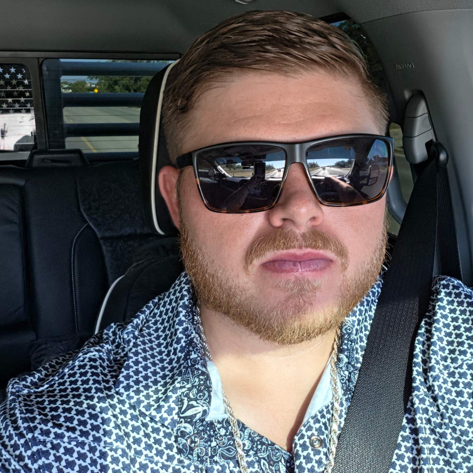 Dylan wearing dark sunglasses and a blue patterned shirt inside a vehicle ready to go